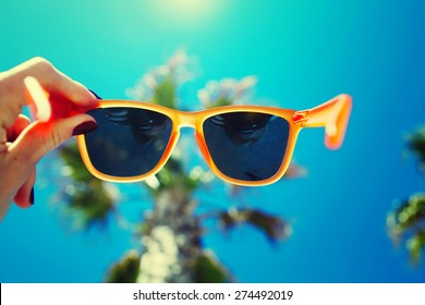 Female Hand Holding Colorful Sunglasses Against Palm Tree And Blue Sunny Sky, Summer Vacation Holidays Concept, First Person Shot, Looking Though Glasses, Filtered Image
