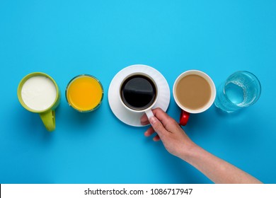 Female Hand Holding A Cup With Black Coffee And Group Of Healthy Drinks, Orange Juice, Cappuccino, Water, Yogurt On A Blue Background. Flat Lay Still Life Table Top View.