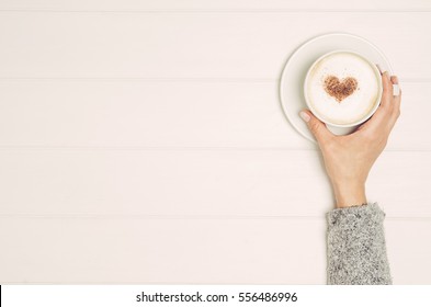 Female Hand Holding Cup Of Coffee On White Wooden Table. Photograph Taken From Above, Top View With Copy Space