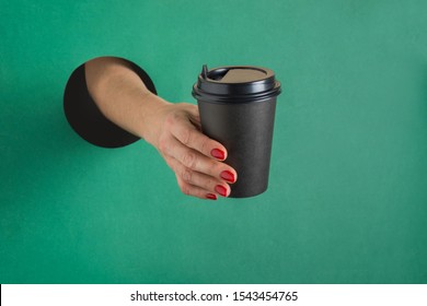 Female Hand Holding Disposable Paper Black Coffee Cup Through Round Hole In Green Paper. Concept