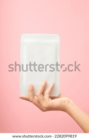 Female hand holding a facial sheet mask white blank realistic mockup on trendy pink background. Space for label design, front view. Daily skincare routine.