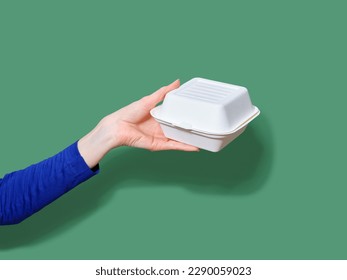 Female hand holding food container for takeaway burger on green background. Flash light, copy space