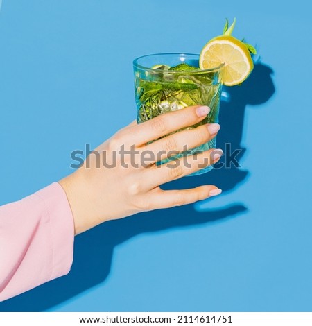 Female hand holding glass with cocktail mojito with lemon isolated on light blue neon background. Complementary colors, blue, yellow and green. Copy space for ad, text, design