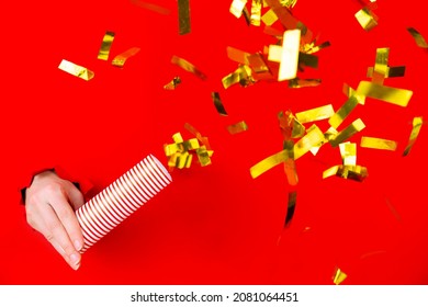 Female Hand Is Holding Golden Confetti Cracker In Torn Hole Of Red Background. Concept Of Winter Holiday, Happy New Year. Merry Christmas. Mockup, Template, Copy Space. Greeting Invitation Card.