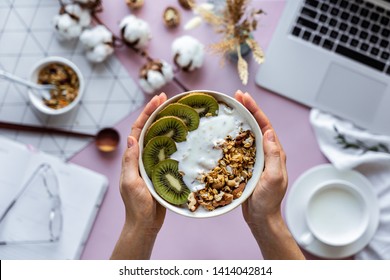 Female hand holding healthy breakfast bowl over work table background concept enjoy detox morning meal with laptop milk, woman eat natural granola nutrition detox food in home office, top view