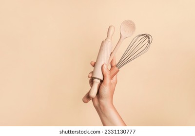 Female hand holding kitchen utensils for food and bakery on beige background. whisk, rolling pin and wooden spoon - Powered by Shutterstock