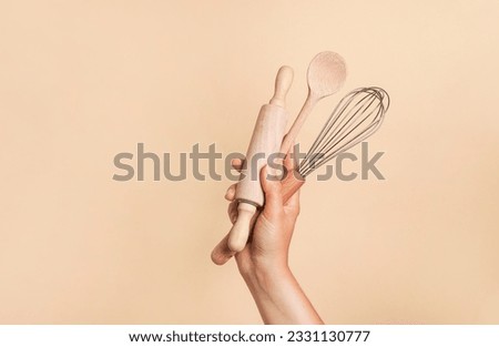 Female hand holding kitchen utensils for food and bakery on beige background. whisk, rolling pin and wooden spoon