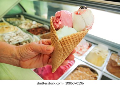 Female Hand Is Holding A Large Strawberry, Mint, Vanilla With Sour Cherry Ice Cream In Waffle Cone. Ice Cream Fridge With Steel Service Containers In Background. Worker In Ice Cream Shop. Pastry Shop.