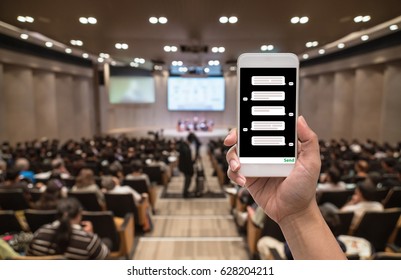 Female Hand Holding Mobile Phone Showing Chat Message When Live Via Social Network Over Abstract Blurred Photo Of Conference Hall Or Seminar Room With Attendee Background, Business Technology Concept