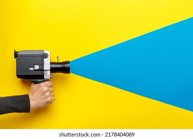 Female Hand Holding Old Style Film Movie Camera On A Yellow Background With Blue Ray Coming Out Of The Camera,  Imitating Shooting Process. Film Making Industry Concept