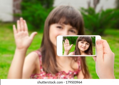 Female Hand Holding A Phone With Video Call Of Little Girl On The Screen On A Background Of Green Grass