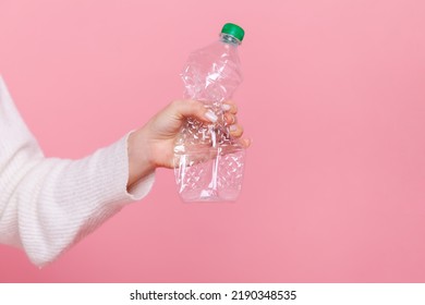 Female hand holding plastic bottle with green cap, sorting her rubbish, worrying about environment, wearing white casual style sweater. Indoor studio shot isolated on pink background.