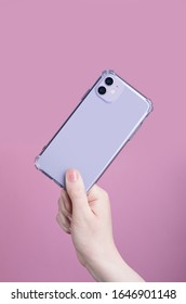 Female Hand Holding Purple IPhone 11 In Transparent Cover Isolated On A Pink Background. Clear Phone Case Mock Up Back View
