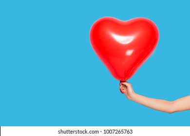 Female Hand Holding A Red Ball In The Shape Of A Heart, Isolated On A Blue Background.
