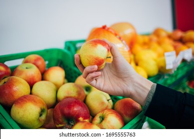 Female Hand Holding Ripe Apple Next To Fruit Shelves In Supermarket, Close-up.