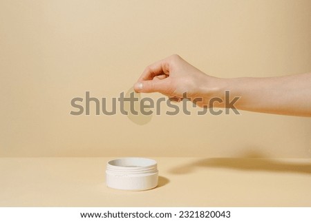 Female hand holding sample of green algae extract eye patch over white jar of product on beige isolated background. The concept of natural product for moisturizing, from dark circles and wrinkles