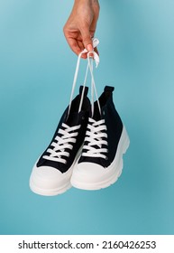 Female hand holding shoelaces stylish and fashionable model of black and white sneakers on blue background. Comfortable black sneakers with white soles and shoelaces for teens or children.