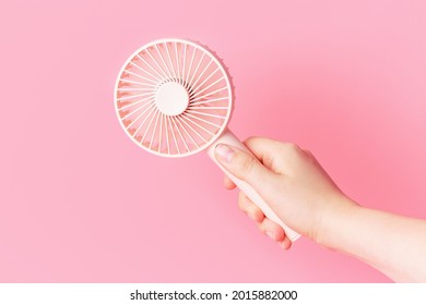 Female hand holding a small fan on pink background close-up.