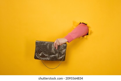 Female Hand Is Holding A Small Golden Clutch Bag With A Metal Chain On A Yellow Background. Part Of The Body Sticking Out Of A Torn Hole In A Paper Background