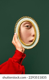 Female hand holding small mirror with reflection of sad woman's face with natural make up over dark green background. Fashion, beauty, creativity, ad. Human emotions, expression concept