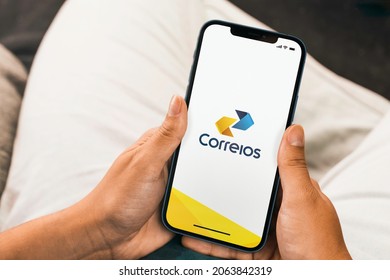 Female Hand Holding A Smartphone With Correios Do Brasil (Post Office Brazil) App On The Screen With A Computer Keyboard. Wooden Background. Rio De Janeiro, RJ, Brazil. October 2021.