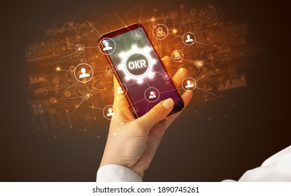 Female hand holding smartphone with OKR abbreviation, modern technology concept