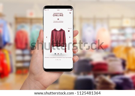 Female hand holding smartphone and shopping online, blurred clothing store in background. Online shopping concept