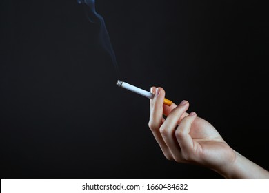 Female Hand Holding A Smoking Cigarette On A Dark Background Copy Space. Harm From Smoking Cigarettes.