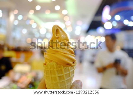 A female hand holding a soft cone of Thai tea ice cream with blur department store area and bokeh light 
