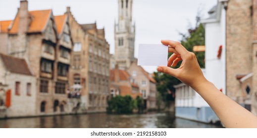 Female hand holding a ticket or business card with copy space on the background of a canal and medieval houses in Bruges, Belgium