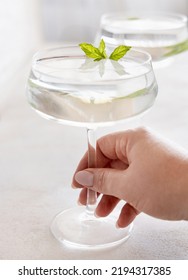 Female Hand Holding Transparent Cocktail In A Coupe Glass Decorated With Mint Leaves Close Up. Tropical Drink At The Bar. Summer Vacation Mood. Analcolic Mocktail Drink