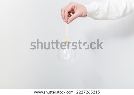 Female hand holding transparent glass Christmas ball on white backdrop. Trendy Christmas background