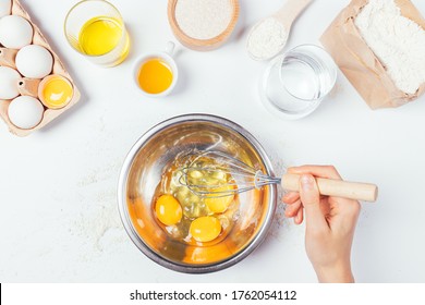 Female hand holding whisk mixes eggs in metal bowl next to ingredients for homemade bakery on white table, top view. - Powered by Shutterstock