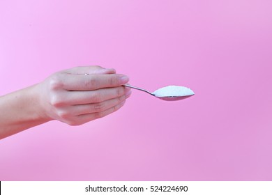 Female Hand Holding White Granulated Sugar In Spoon On Pink Background