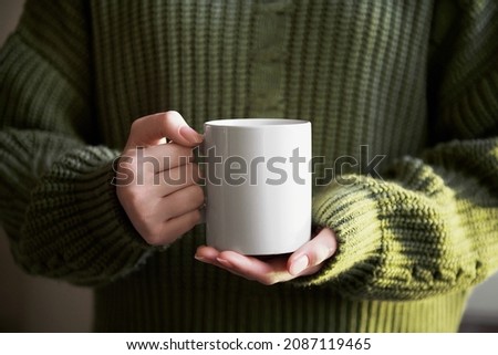 female hand holding white mug with blank copy space for your advertising text message or promotional content, sweet coffee or tea. Girl in green sweater holding white porcelain mug mock up