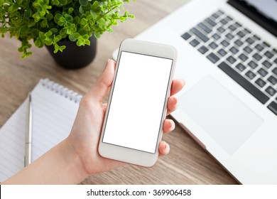 Female Hand Holding A White Phone With Isolated Screen On A Table With Laptop