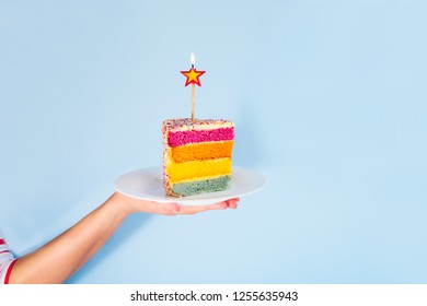 Female Hand Holding White Plate With Slice Of Rainbow Cake With Birning Candle In The Shape Of Star Isolated On Blue Background. Happy Bithday, Party Concept. Square Card. Selective Focus. Copy Space