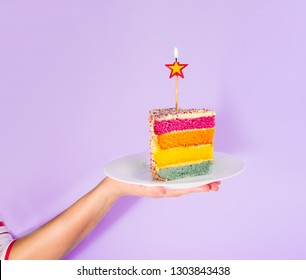 Female Hand Holding White Plate With Slice Of Rainbow Cake With Birning Candle In The Shape Of Star Isolated On Blue Background. Happy Bithday, Party Concept. Square Card. Selective Focus. Copy Space