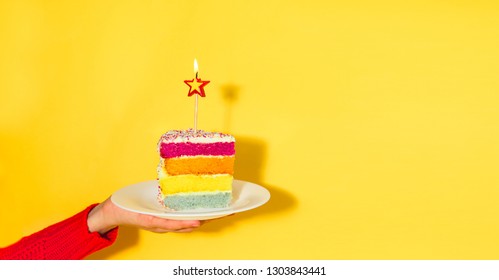 Female Hand Holding White Plate With Slice Of Rainbow Cake With Birning Candle In The Shape Of Star Isolated On Yellow Background. Happy Bithday, Party Concept. Wide Banner. Copy Space