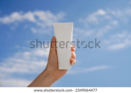 Female hand holding a white wet mockup jar of body or face cream on a blue sky background with clouds. Concept of cosmetics with spf, moisturizing and skin protection