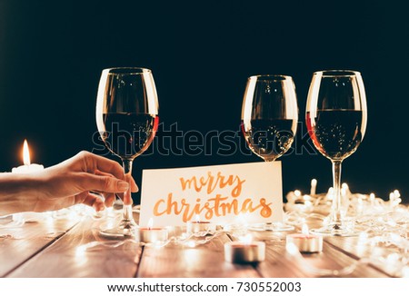Female hand holding a wine glass over wooden table with fairylights, candles and merry christmas card