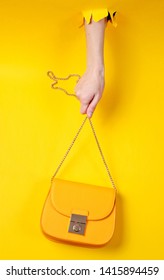 Female Hand Is Holding Yellow Leather Bag Over Chain Through Torn Yellow Paper. Minimalistic Fashion Concept