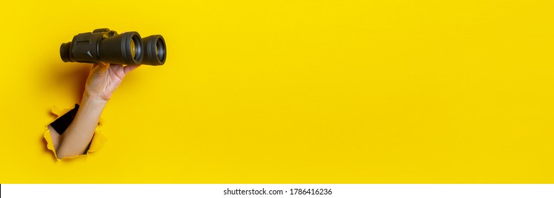 Female hand holds black binoculars on yellow background, travel, find and search concept. Banner.