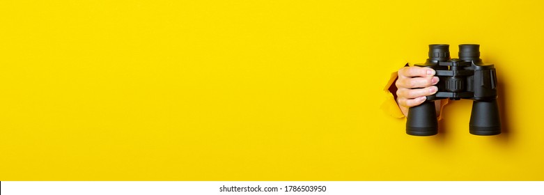 Female hand holds black binoculars on a yellow background. Looking through binoculars, travel, find and search concept. Banner.