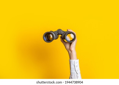 female hand holds black binoculars on a bright yellow background.
