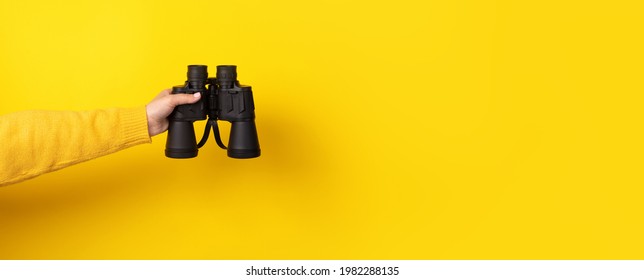 Female hand holds black binoculars on a yellow background. Looking through binoculars, travel, find and search concept. Banner. - Shutterstock ID 1982288135