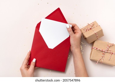 Female hand holds blank square paper mockup envelope red, golden stars confetti, gift boxes on beige background. Flat lay, top view, copy space, minimalist. Christmas and New Year concept - Powered by Shutterstock