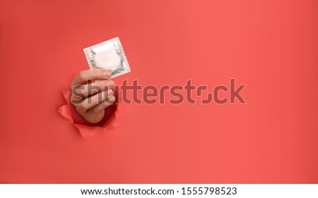 Female hand holds condom on torn red paper wall. Safe pleasure and protection, contraception, protection from AIDS and day to fight AIDS. Copy space aside for your advertising content