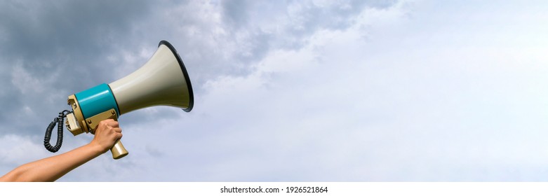 A female hand holds a megaphone. Horn loudspeaker is big and old. Clouds background. The concept of a message in business, loudspeaker, attracting attention.