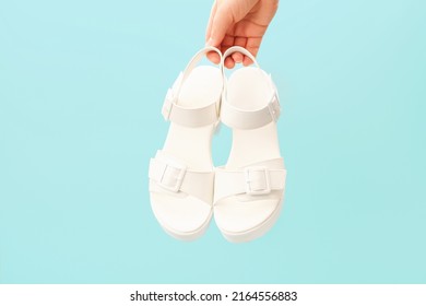 Female Hand Holds Pair Of White Sandals On Blue Background Close-up, Concept Of Summer Shoes.